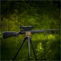 Trójnóg Primos Trigger Stick Apex - zdjęcie karabinu opartego na trójnogu wśród zielonej roślinności