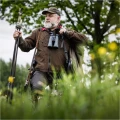 Trójnóg Primos Trigger Stick Gen III™ 24-62'' - zdjęcie w plenerze, myśliwy z lornetką idący przez wysoką trawę, z trójnogiem w dłoni
