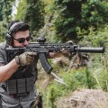 Celownik pryzmatyczny Primary Arms GLx 1x Micro Prism iR Red ACSS Cyclops Gen III -myśliwy celujący z broni z celownikiem na rozmytym tle lasu