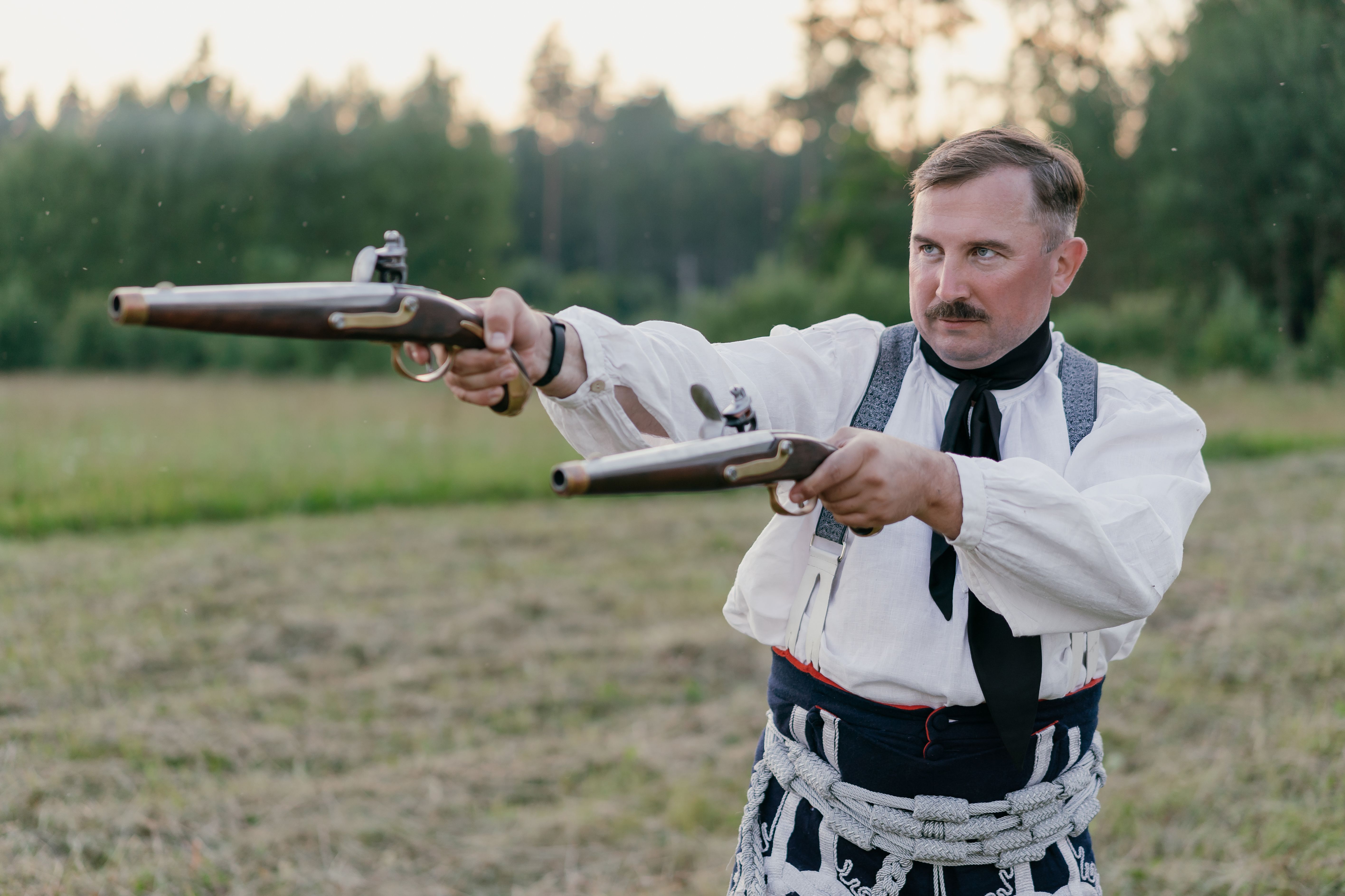 Strzelec w stroju historycznym z dwoma pistoletami skałkowymi na łące, w trakcie strzału